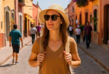 Mujer joven con sombrero de turista y lentes oscuros caminando por una calle soleada de Tequisquiapan, Querétaro, rodeada de personas y arquitectura colonial.
